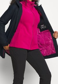 Femme portant une veste polaire rose vif et un pantalon noir ouvre un manteau noir, révélant une doublure matelassée rose et le logo CMP à l'intérieur.