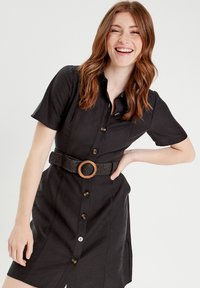 Robe noire à manches courtes avec un bouton sur le devant, taille cintrée mettant en valeur une ceinture tissée avec une boucle en bois, et un ourlet droit.