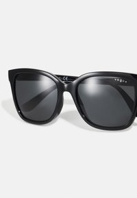 Ochelari de soare din plastic negru, cu lentile mari pătrate și finisaj mat. Prezintă un logo pe braț și lentile fumurii închise.