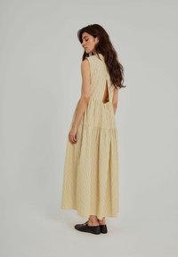 Gulstribet maxi kjole med høj hals og åben ryg. Lagdelt skirt når anklerne. Modellen bærer brune flade sko.
