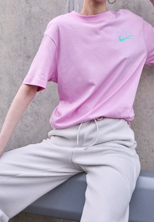 Personne portant un t-shirt rose Nike et un pantalon de jogging en polaire blanc, assise contre un mur en béton avec le bras tendu.