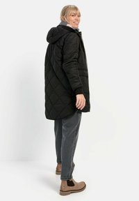 Schwarzer gesteppter langer Mantel mit Kapuze, gerippten Bündchen und silbernem Reißverschluss. Kombiniert mit grauen Jeans und braunen Stiefeletten mit elastischen Seiteneinsätzen.