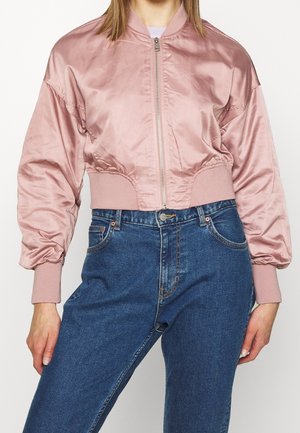 Nainen, joka käyttää kiiltävää pölyisen vaaleanpunaista lyhennettyä bomber-takkia, jossa on resorit hihansuissa, ja korkeavyötäröisiä sinisiä farkkuja valkoista taustaa vasten.