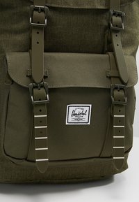 Oliwkowy plecak z teksturowanej tkaniny, z regulowanymi paskami oraz przednią klapą z naszywką logo i zapięciami na paski.