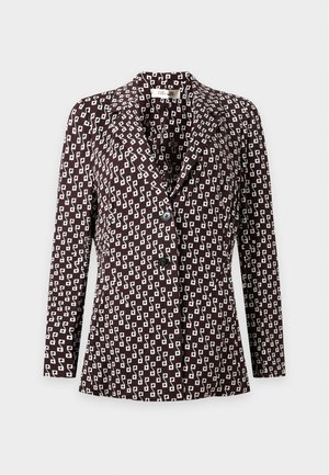 Femme debout, portant un tailleur-pantalon foncé avec un motif géométrique clair, tenant un petit sac noir avec une chaîne dorée et des chaussures noires pointues.