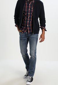 Giacca a zip trapuntata blu navy sopra una camicia a quadri rossa e blu navy, abbinata a jeans azzurri chiari e sneakers bianco e nero.