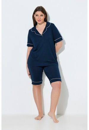 Frau steht barfuß und trägt ein marineblaues kurzärmeliges Oberteil sowie kniehohe Shorts mit weißen Paspelierungen am Kragen und Saum.