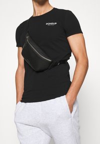 Svart läderbälte väska med dragkedja och justerbar rem bärs över en svart t-shirt. Matchar med ljusgrå svettbyxor.