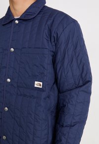 Isolerad marinblå jacka med ett kviltat mönster, tryckknappar och en bröstficka med en logotag. Mjuk textur och krage.