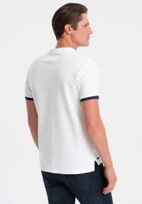 Bílé polo tričko s krátkými rukávy, tmavě modré detaily na manžetách, žebrovaná textura, rovný lem a boční rozparky pro větší pohodlí.