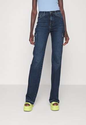 Dunkelblaue Denim-Cargohosen mit geraden Beinen, zwei Seitentaschen und sichtbaren Nähten. Kombiniert mit einem hellblauen Oberteil und grünen Plateauschuhen.