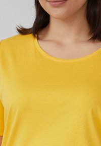 Frau trägt ein hellgelbes Rundhals-T-Shirt, das einen Teil ihres Halses, Kinns und schulterlangen dunklen Haars zeigt.