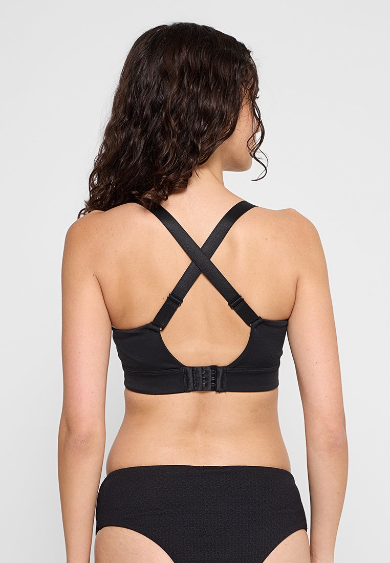 Kobieta z długimi, kręconymi włosami, ubrana w czarny biustonosz z krzyżującymi się ramiączkami oraz czarne, strukturalne majtki, pokazana z tyłu na jednolitym szarym tle.