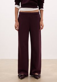 Pantaloni larghi bordeaux in felpa realizzati in un tessuto morbido, con vita elastica e superficie liscia e priva di decorazioni. Abbinati a ballerine scure decorate con un fiocco.