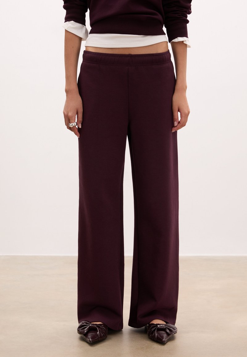 Pantaloni larghi bordeaux in felpa realizzati in un tessuto morbido, con vita elastica e superficie liscia e priva di decorazioni. Abbinati a ballerine scure decorate con un fiocco.