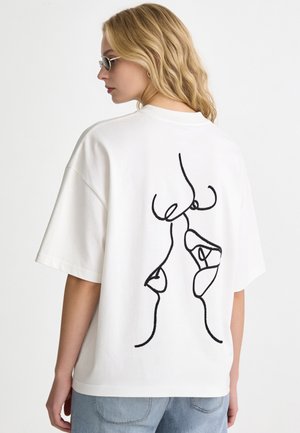 Donna bionda con occhiali da sole e una t-shirt bianca oversize con un disegno astratto a linee nere sulla schiena.
