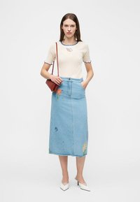 Γυναίκα που στέκεται φορώντας μπεζ τοπ με κοντά μανίκια και μπλε διακοσμητική επένδυση, μπλε τζιν μίντι φούστα με κεντήματα από λουλούδια, λευκά τακούνια και καφέ τσάντα ώμου.