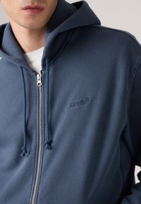 Nærbilde av person som har på seg en glidelås navy blå Levi's hettegenser over en hvit skjorte, som viser snorer og brodert logo på brystet.