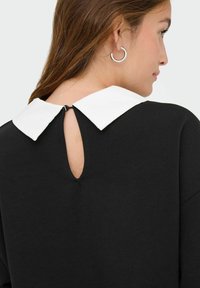 Abito nero con colletto bianco; presenta un'apertura a forma di chiave sul retro. Il tessuto sembra liscio con una vestibilità comoda. Orecchino a cerchio in argento visibile.