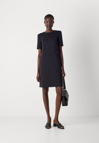 Robe noire à manches courtes, coupe droite, avec des poches latérales et de subtils plis aux épaules. Associée à des sandales noires à enfiler et un sac à main.