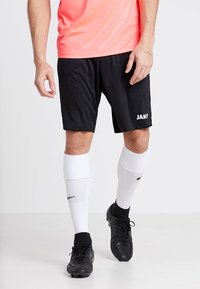 Juodi sportiniai šortai su "JAKO" logotipu, derinami su baltais kelių ilgio kojiniais ir juodais futbolo batais. Viršus yra ryškiai rožinės spalvos, tekstūruoto audinio.