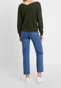 Grüner Strickpullover mit schulterfreiem Ausschnitt, kombiniert mit blauen geraden Jeans und weißen Turnschuhen, zeigt eine gerippte Textur und eine lockere Passform.