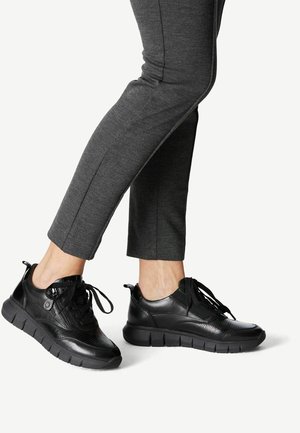 Personne portant un pantalon ajusté gris foncé et des baskets en cuir noir à semelles épaisses et lacets, debout avec un pied légèrement levé.