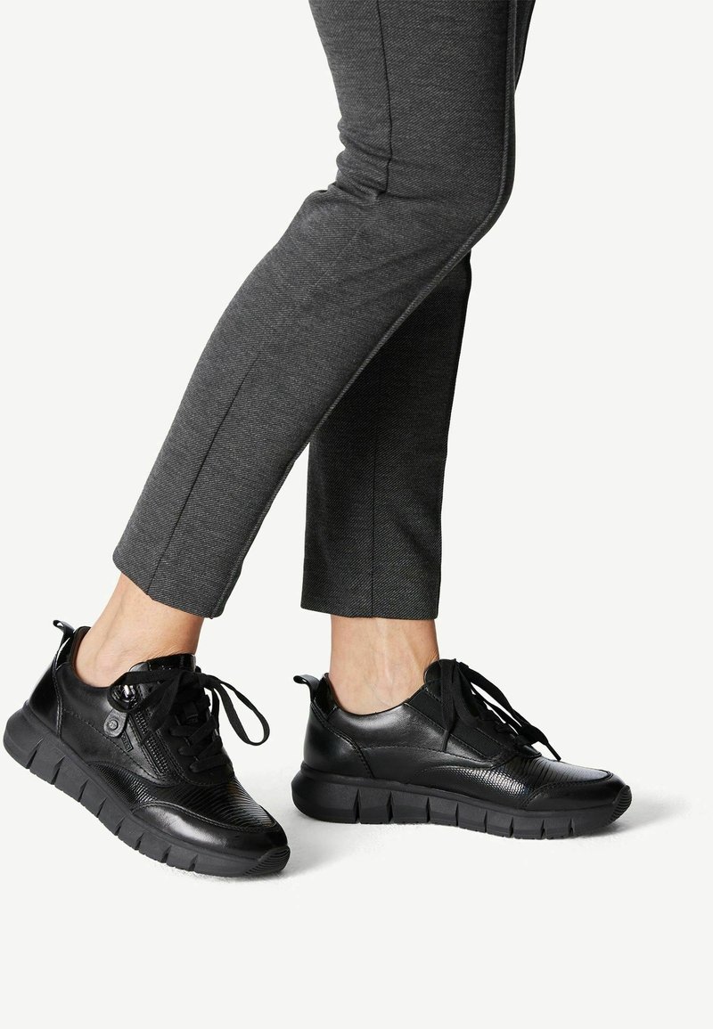 Personne portant un pantalon ajusté gris foncé et des baskets en cuir noir à semelles épaisses et lacets, debout avec un pied légèrement levé.