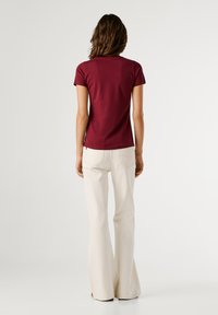 Žena s vlnitými hnedými vlasmi, oblečená v priliehavom burgundskom tričku s krátkym rukávom a bielych nohaviciach s vysokým pásom, stojí chrbtom.