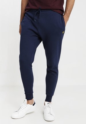 Man wearing navy blue joggers with elastic cuffs and white sneakers, standing against a plain light background.