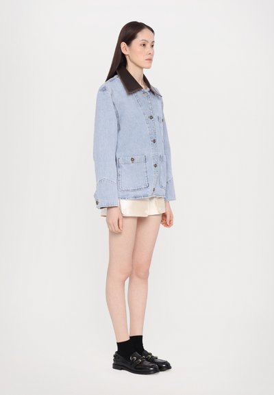 Lichtblauwe spijkerjack met een donkerbruine kraag, voorzien van knopen en twee voorzakken, gedragen over een crèmekleurige rok en zwarte schoenen.