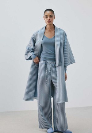 Vrouw die blauwe gestreepte wijd uitlopende broek en een bijpassende lange jas draagt over een aangesloten blauwe V-hals top, staande tegen een effen lichte achtergrond.