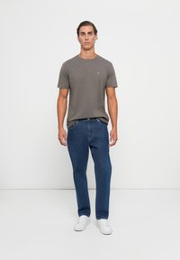 Ung mann står mot en hvit bakgrunn iført en brun t-skjorte, blå jeans og hvite sneakers.