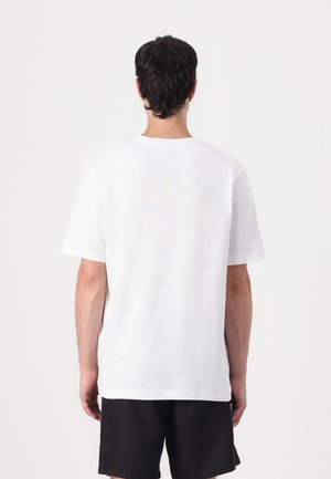 Homme aux cheveux foncés debout de dos, portant un t-shirt blanc uni à manches courtes et un short noir, sur un fond blanc.