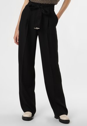 Persoon draagt een zwarte broek met wijde pijpen en strikriem en witte schoenen, staat met de handen in de zakken tegen een effen achtergrond.