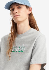 Camiseta de algodón gris con el logo bordado de 'Levi's' en verde y blanco. Combinada con una gorra de mezclilla azul clara. Cabello largo y liso.