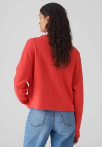 Pull rouge à col rond avec une coupe décontractée, des manches longues et un ourlet droit. Porté avec un jean taille haute bleu clair, mettant en valeur le design arrière.