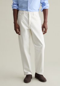 Pantalones de mezclilla blancos con un corte recto, que cuentan con cremallera en la entrepierna, bolsillos frontales y detalles de costura sutiles, combinados con zapatos marrones.