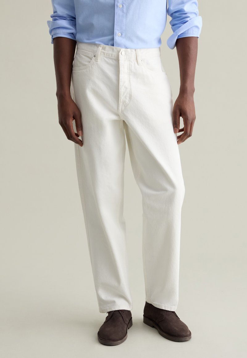 Pantalones de mezclilla blancos con un corte recto, que cuentan con cremallera en la entrepierna, bolsillos frontales y detalles de costura sutiles, combinados con zapatos marrones.