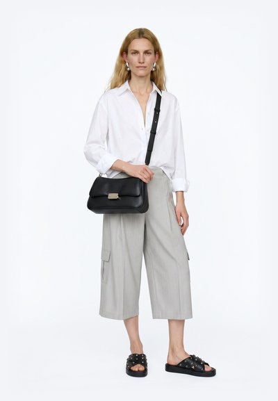 Woman wearing white shirt and light gray wide-leg cropped pants, holding a black shoulder bag and wearing black studded slide sandals.