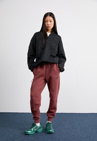 Sweatshirt preto com fecho, com padrão texturizado, calças de jogging bordô com bolsos laterais e ténis verdes desportivos com detalhes reflexivos.