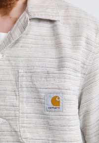 Leichtes, gestreiftes Hemd in Off-White, mit einer Brusttasche und einem Carhartt-Logopatch in Gelb und Grau. Horizontale Textur.