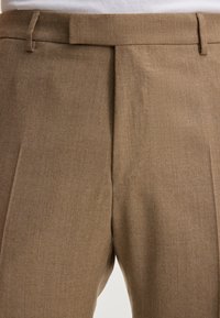 Pantaloni beige su misura realizzati in tessuto texturizzato, con un frontale piatto, due tasche laterali e passanti per la cintura in vita.