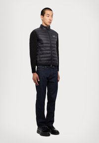 Gilet matelassé noir avec col haut et fermeture éclair à l'avant, porté sur une chemise à manches longues noire à motifs. Un jean sombre et des chaussures noires à lacets complètent la tenue.
