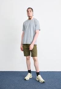 T-shirt gris clair à manches courtes associé à un short vert olive et des baskets jaunes. Le modèle se tient sur un sol moquetté bleu contre un mur blanc.