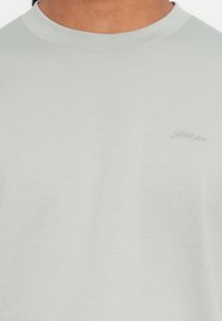 Άτομο που φορά ελαφρώς γκρι μπλουζάκι με στρογγυλή λαιμόκοψη, με το μικρό, διακριτικό λογότυπο "Hollister" κεντημένο στο αριστερό στήθος.