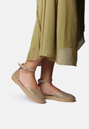 Feet wearing beige espadrille flats with ankle straps paired with a light green flowing skirt against a white background.