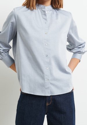 Kobieta w jasnoniebieskiej koszuli zapinanej na guziki z podwiniętymi długimi rękawami, w ciemnoniebieskich dżinsach, ręce schowane za plecami.