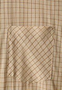 Tissu à carreaux beige et marron avec une poche carrée cousue, présentant des lignes fines sombres et claires qui se croisent en motif quadrillé.