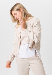 Beige cropped jack met drukknoopjes en borstzakken, gedragen over een witte top. Glad textiel met een lichte val, gecombineerd met bijpassende beige broeken.